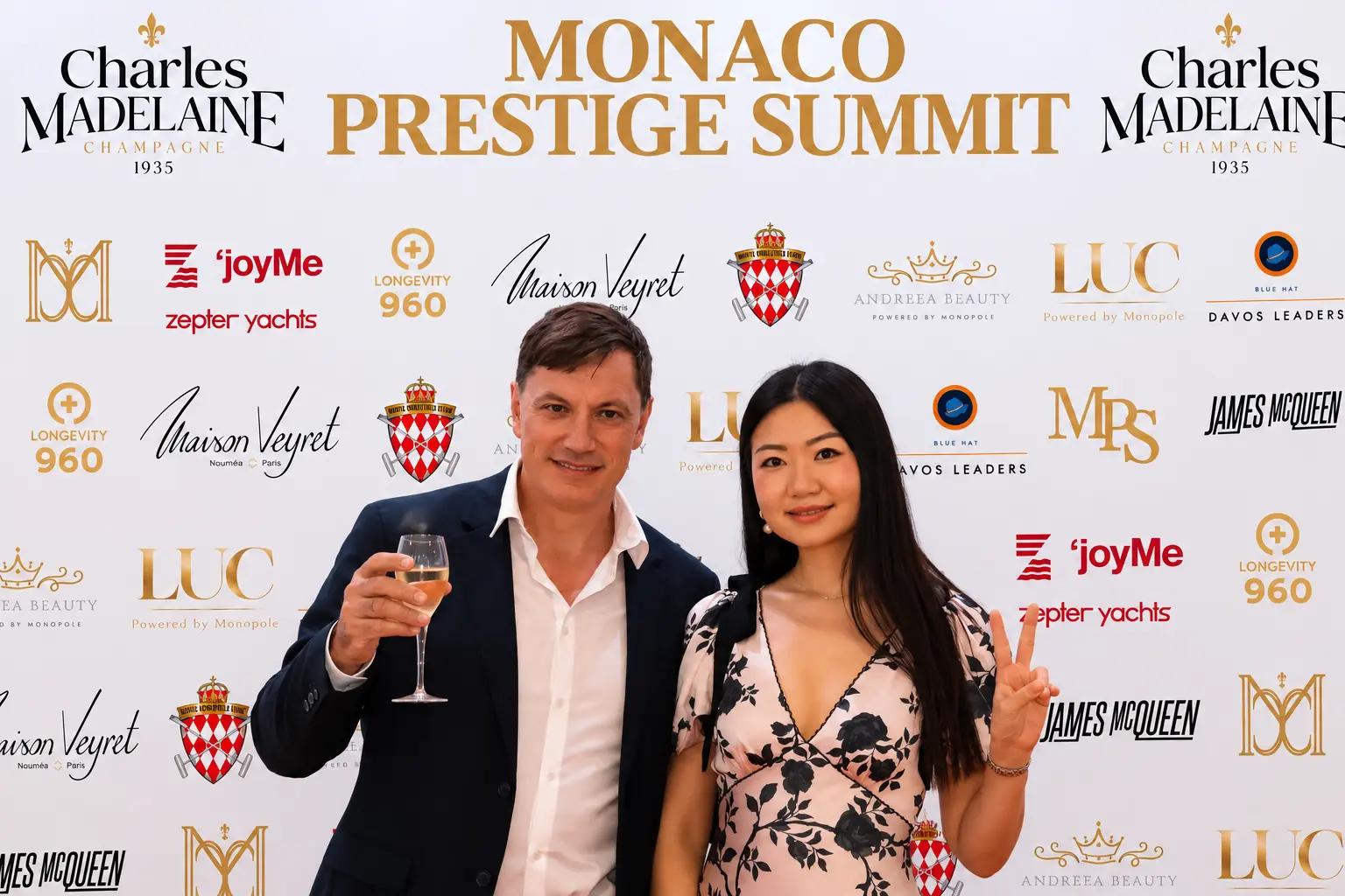 Guest toasting champagne beside a guest in a floral dress at the Monaco Prestige Summit media wall